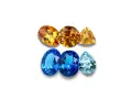 A collection of faceted gemstones, including warm orange and yellow topaz birthstones, and cool blue and aquamarine stones, arranged in two rows.