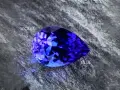 A pear-shaped tanzanite gemstone, displaying vibrant blue and violet hues, rests on a textured grey surface. Is tanzanite expensive? This gem's rarity suggests yes.