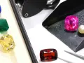 A blue sapphire, green emerald, and yellow citrine are displayed in a box, while a pink sapphire, pearl, and red garnet are on a tray, showcasing diverse gemstone families.