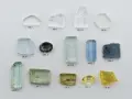A collection of raw and cut gemstones, including a yellow heliodor gemstone, aquamarines, and other beryl varieties, displayed on a white background.