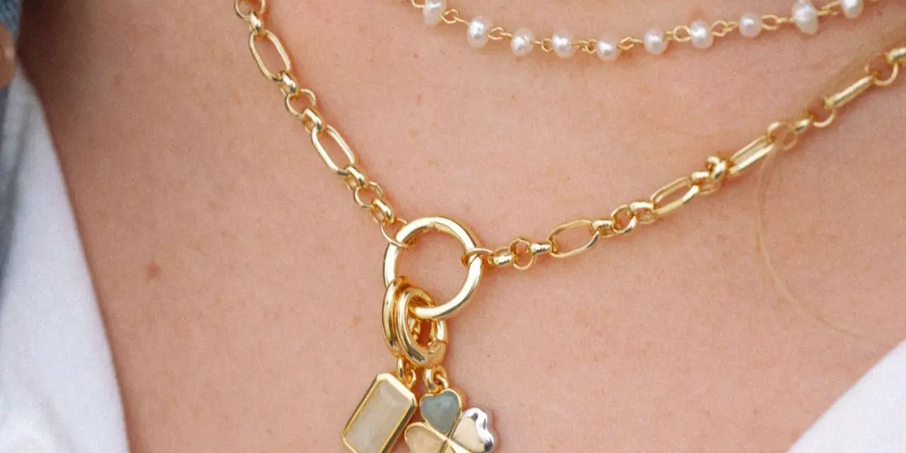Layered necklaces featuring different types of gold chains, including a figaro chain and a pearl strand, adorned with charms.