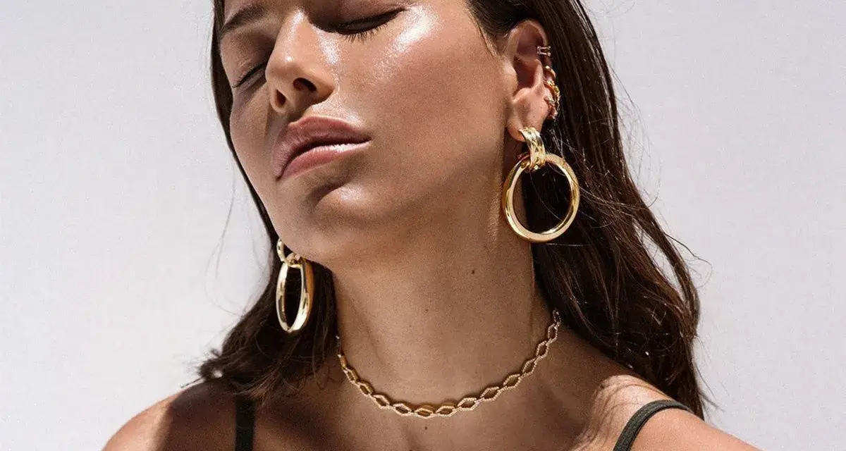 A woman with closed eyes showcases various types of dangling earrings, including large gold hoops and smaller ear cuffs.