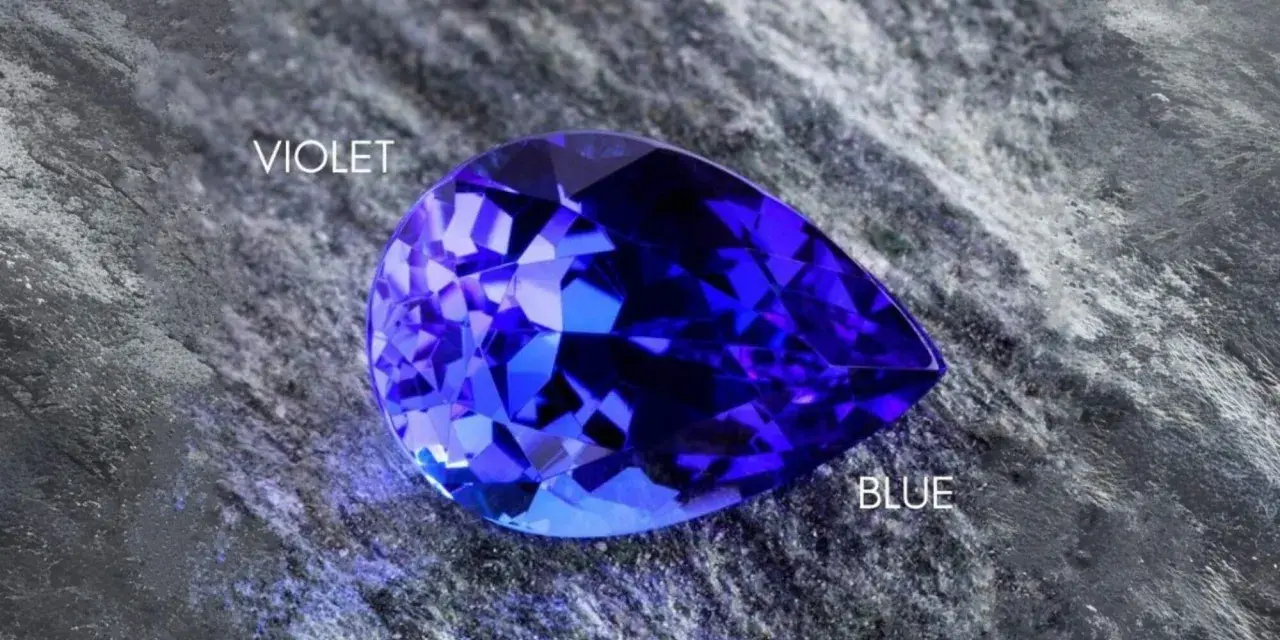 A pear-shaped tanzanite gemstone, displaying vibrant blue and violet hues, rests on a textured grey surface. Is tanzanite expensive? This gem's rarity suggests yes.