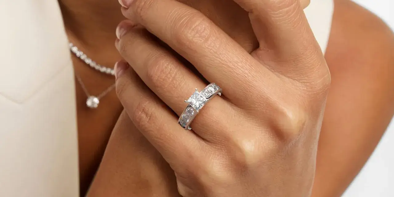 A woman's hand displays a stunning ring featuring a square princess cut diamond, flanked by smaller stones. A delicate diamond necklace is also visible.