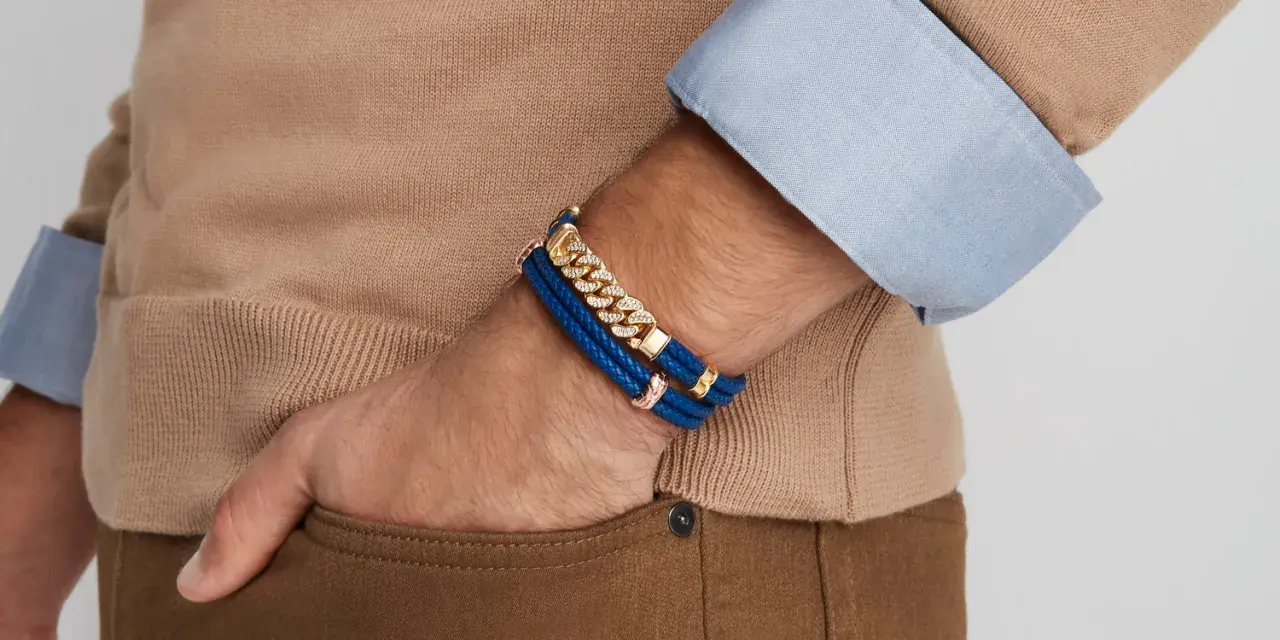 A man's wrist displays two stylish bracelet types: a blue braided leather bracelet with gold accents and a diamond-studded gold chain bracelet.