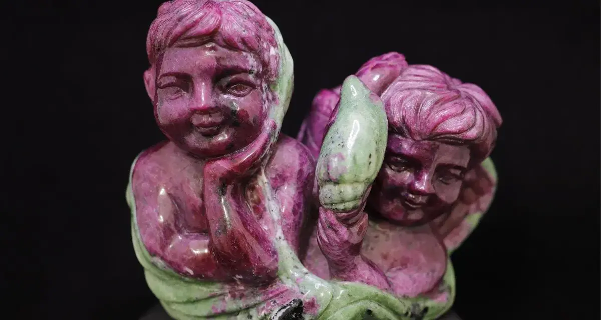 Two cherubic figures carved from ruby in zoisite, one resting its head on its hand, the other holding a green leaf-like element.