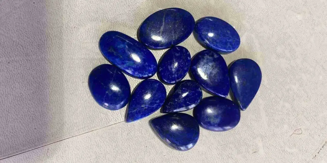 A collection of polished, deep blue Afghanistan gemstones, likely lapis lazuli, are scattered on a textured surface.