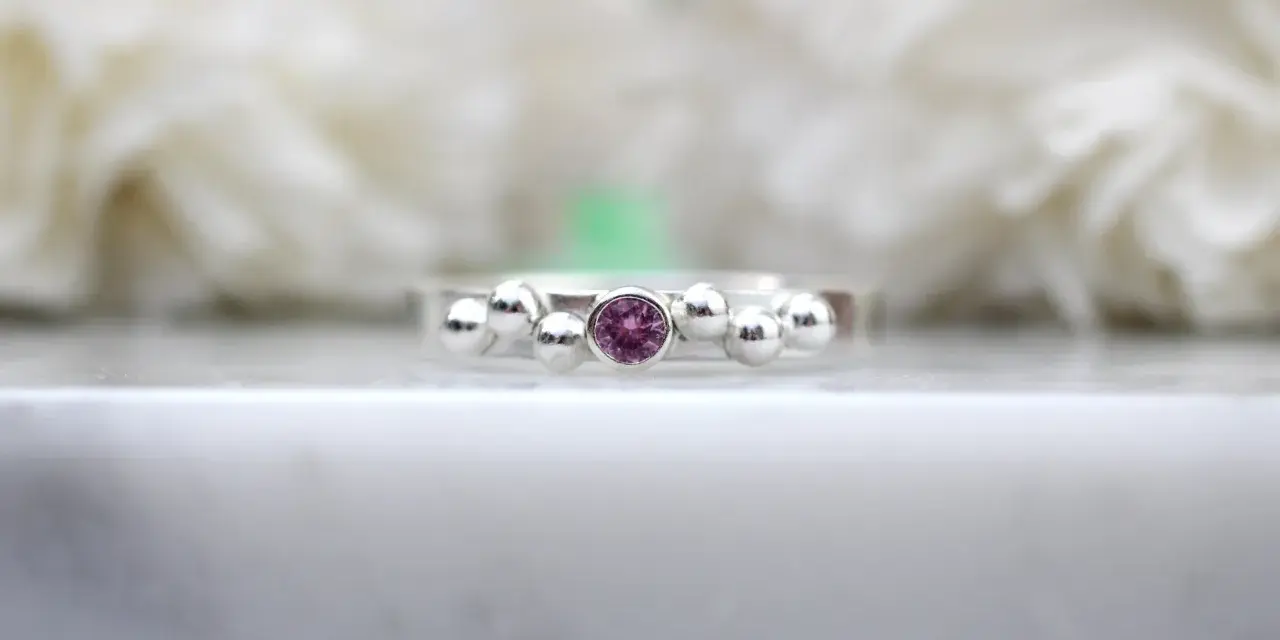 A silver ring with a central pink sapphire, the birthstone for September, surrounded by small silver spheres.