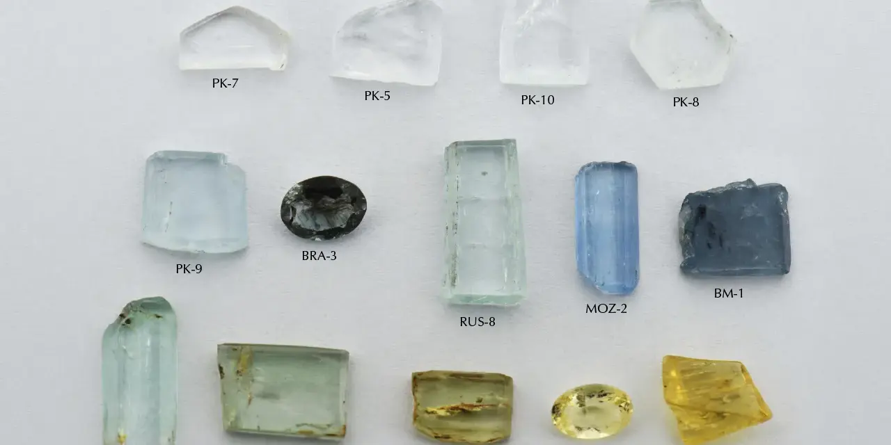 A collection of raw and cut gemstones, including a yellow heliodor gemstone, aquamarines, and other beryl varieties, displayed on a white background.