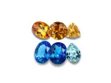A collection of faceted gemstones, including warm orange and yellow topaz birthstones, and cool blue and aquamarine stones, arranged in two rows.