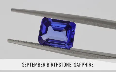 A faceted, deep blue sapphire, the September birthstone, is held by silver tweezers against a light gray background.
