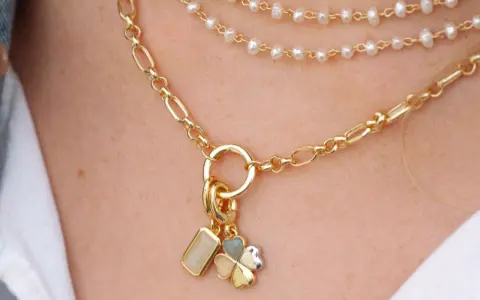 Layered necklaces featuring different types of gold chains, including a figaro chain and a pearl strand, adorned with charms.