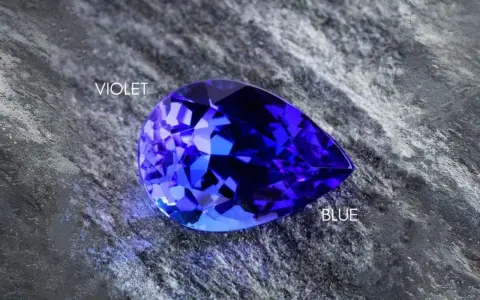 A pear-shaped tanzanite gemstone, displaying vibrant blue and violet hues, rests on a textured grey surface. Is tanzanite expensive? This gem's rarity suggests yes.
