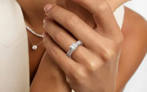 A woman's hand displays a stunning ring featuring a square princess cut diamond, flanked by smaller stones. A delicate diamond necklace is also visible.