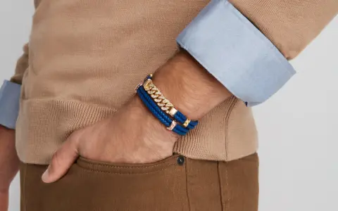 A man's wrist displays two stylish bracelet types: a blue braided leather bracelet with gold accents and a diamond-studded gold chain bracelet.