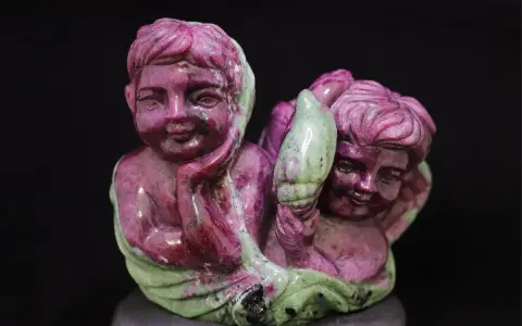 Two cherubic figures carved from ruby in zoisite, one resting its head on its hand, the other holding a green leaf-like element.