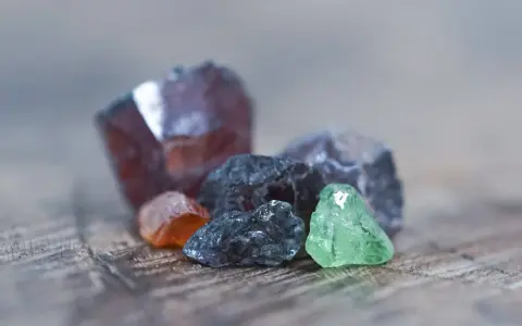 Raw gemstones in various colors, including deep red, orange, grey, and green. The deep red stone might be the birthstone for January.