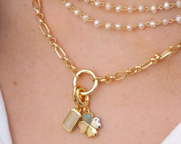 Layered necklaces featuring different types of gold chains, including a figaro chain and a pearl strand, adorned with charms.
