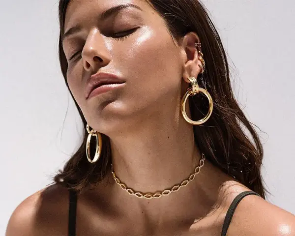 A woman with closed eyes showcases various types of dangling earrings, including large gold hoops and smaller ear cuffs.