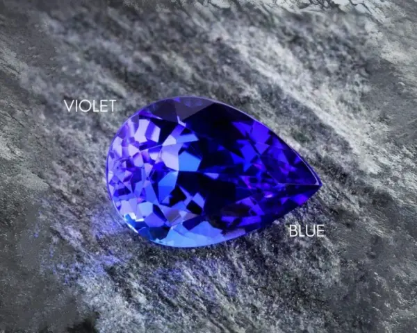 A pear-shaped tanzanite gemstone, displaying vibrant blue and violet hues, rests on a textured grey surface. Is tanzanite expensive? This gem's rarity suggests yes.