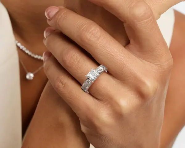 A woman's hand displays a stunning ring featuring a square princess cut diamond, flanked by smaller stones. A delicate diamond necklace is also visible.