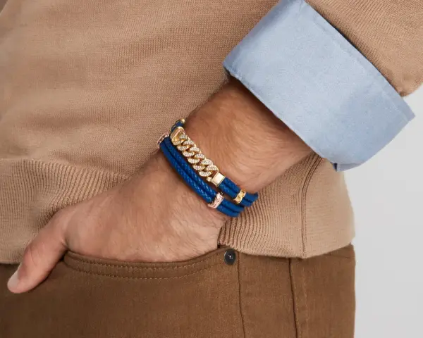 A man's wrist displays two stylish bracelet types: a blue braided leather bracelet with gold accents and a diamond-studded gold chain bracelet.