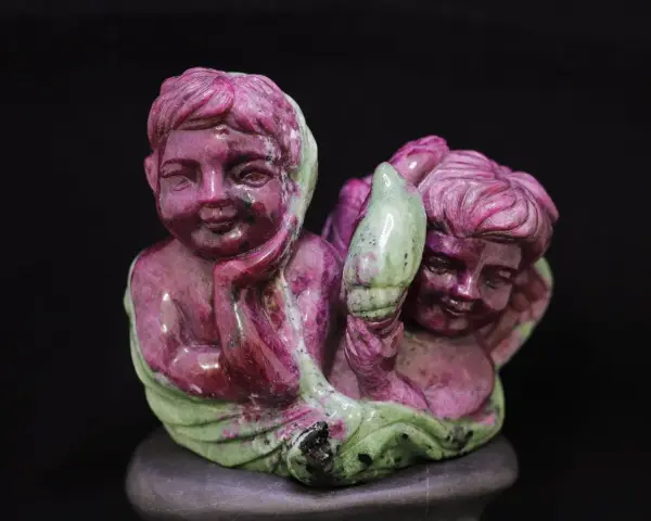 Two cherubic figures carved from ruby in zoisite, one resting its head on its hand, the other holding a green leaf-like element.