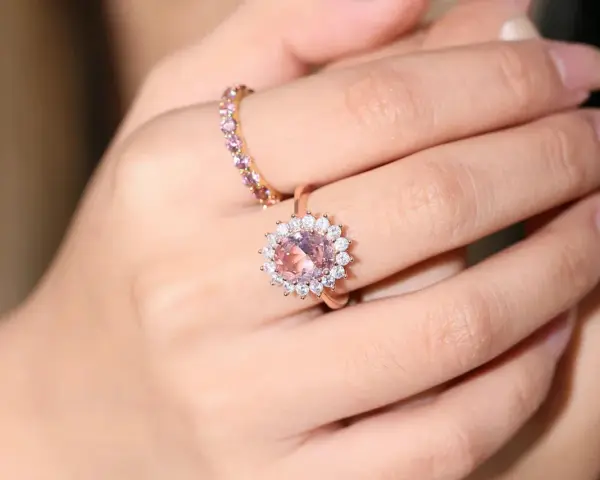 A hand wears two rings. The main ring features a large, peachy-pink morganite surrounded by small diamonds.