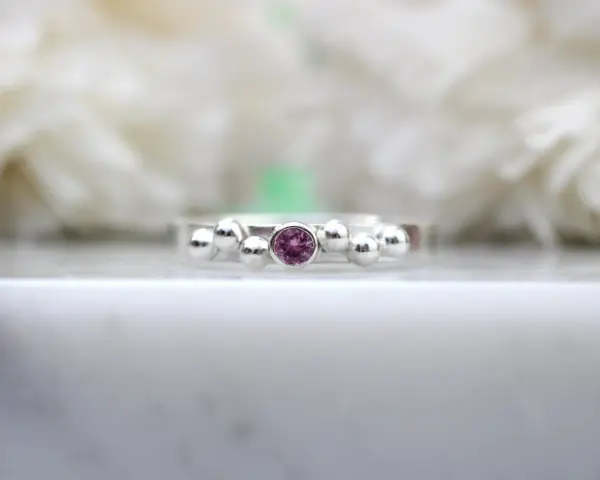 A silver ring with a central pink sapphire, the birthstone for September, surrounded by small silver spheres.