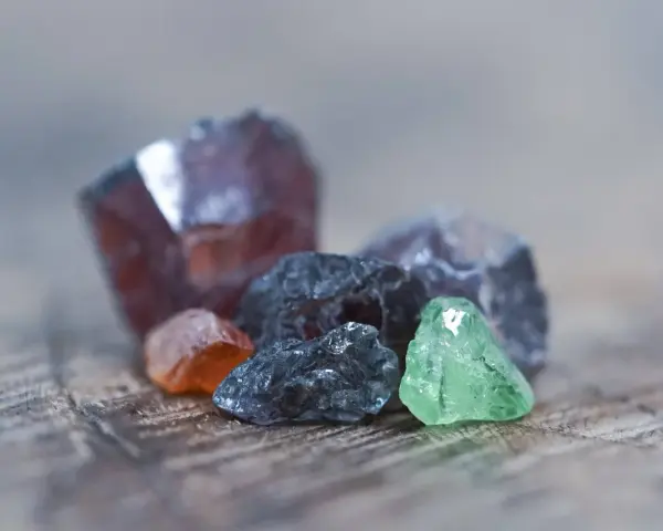 Raw gemstones in various colors, including deep red, orange, grey, and green. The deep red stone might be the birthstone for January.