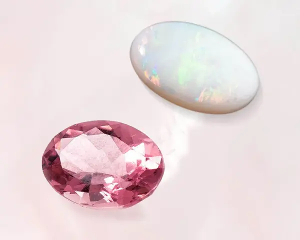 Two oval gemstones, a pink tourmaline and a white opal, are displayed. The opal's play-of-color makes it a perfect october 27 birthstone.