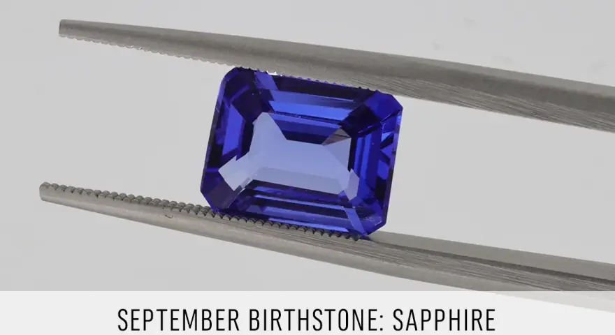 A faceted, deep blue sapphire, the September birthstone, is held by silver tweezers against a light gray background.