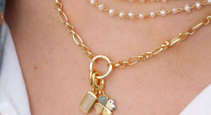 Layered necklaces featuring different types of gold chains, including a figaro chain and a pearl strand, adorned with charms.