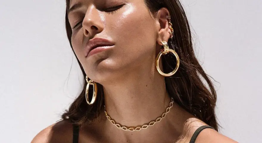A woman with closed eyes showcases various types of dangling earrings, including large gold hoops and smaller ear cuffs.