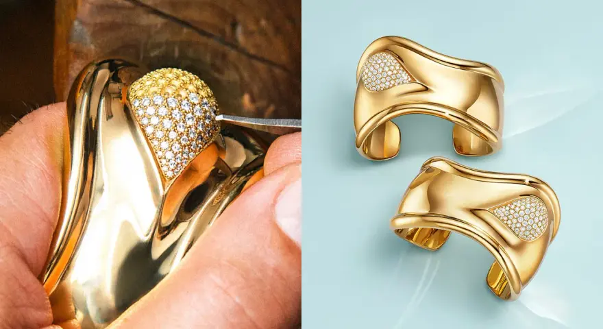 A jeweler meticulously sets diamonds on a gold ring, a testament to Elsa Peretti's exquisite designs. Two finished gold cuffs with diamond accents gleam on a light blue background.