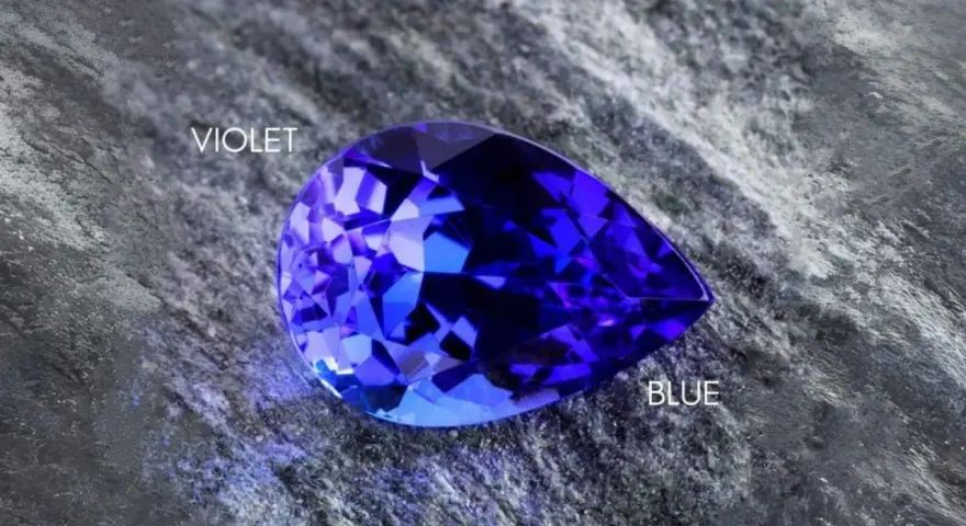 A pear-shaped tanzanite gemstone, displaying vibrant blue and violet hues, rests on a textured grey surface. Is tanzanite expensive? This gem's rarity suggests yes.