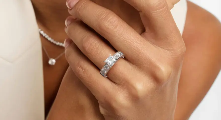 A woman's hand displays a stunning ring featuring a square princess cut diamond, flanked by smaller stones. A delicate diamond necklace is also visible.