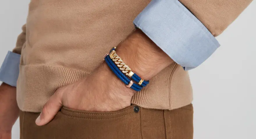 A man's wrist displays two stylish bracelet types: a blue braided leather bracelet with gold accents and a diamond-studded gold chain bracelet.