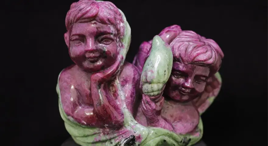 Two cherubic figures carved from ruby in zoisite, one resting its head on its hand, the other holding a green leaf-like element.
