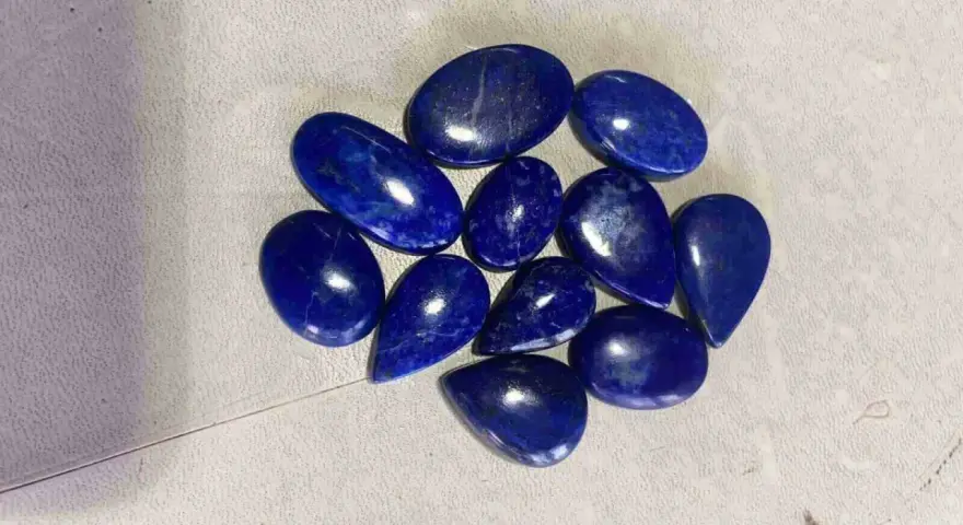 A collection of polished, deep blue Afghanistan gemstones, likely lapis lazuli, are scattered on a textured surface.