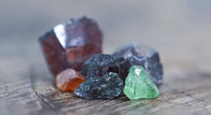 Raw gemstones in various colors, including deep red, orange, grey, and green. The deep red stone might be the birthstone for January.