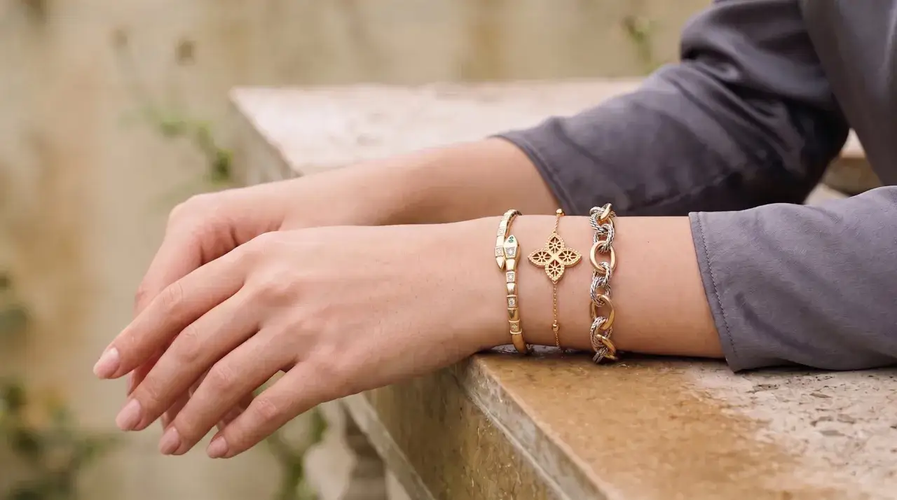 A collection of elegant bracelet types adorns a wrist: a diamond-studded snake, a gold clover charm, and a two-tone chain.