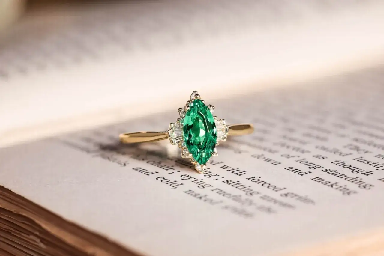 A marquise-cut emerald ring, the perfect May 4th birthstone, rests on an open book, its green gem sparkling.