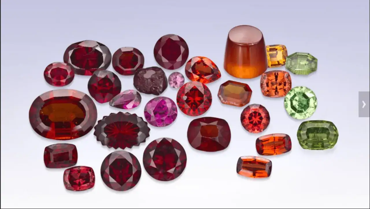 A collection of faceted gemstones in various garnet colors, from deep red to orange and green, arranged on a light blue background.