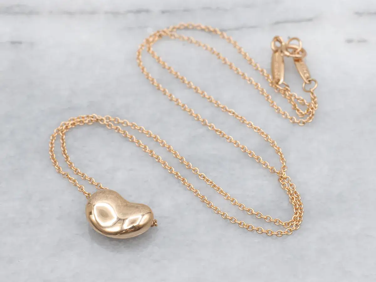 A delicate gold necklace with a heart-shaped pendant, a classic Elsa Peretti design, rests on a marble surface.