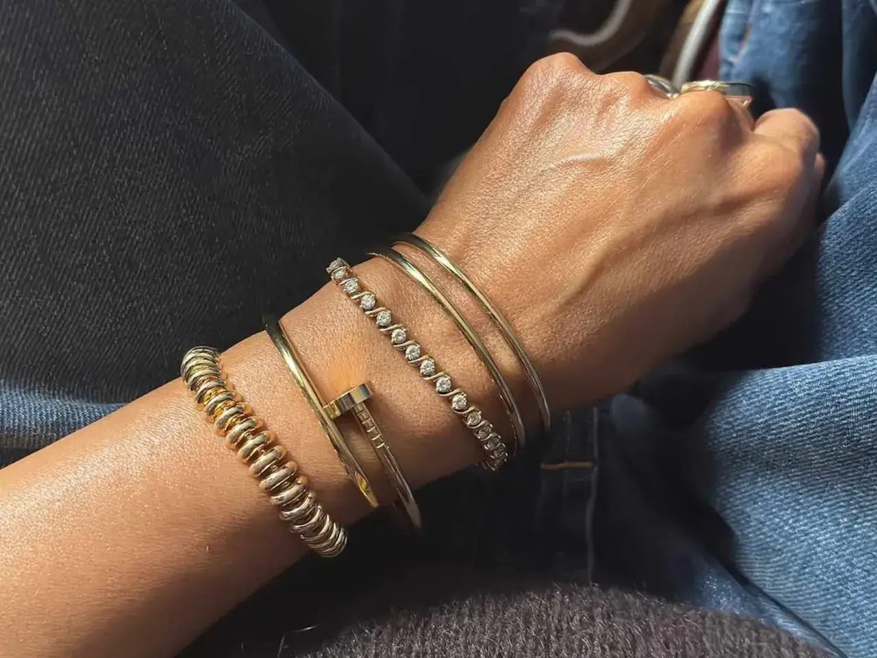 A hand adorned with a stack of gold jewelry, including a diamond bangle and a nail-shaped bracelet, showcasing a chic bangle vs bracelet style.