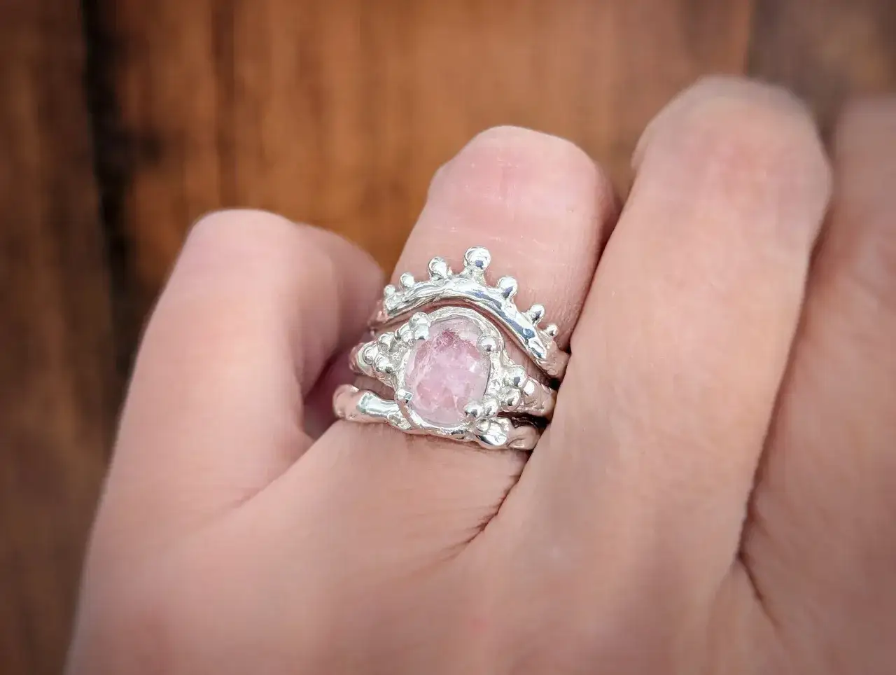 A stack of silver rings on a finger, featuring a raw, oval-cut pink gemstone. This beautiful stone is what's morganite, known for its delicate blush hue.