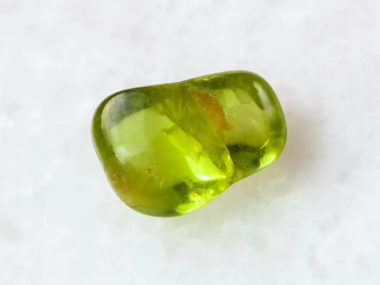 A smooth, translucent peridot stone, known for its vibrant green hue and peridot stone meaning of renewal and growth, rests on a white marble surface.