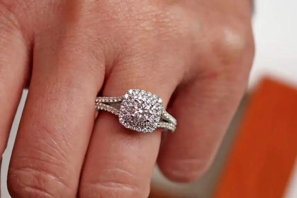 A hand wears a sparkling engagement ring featuring a central composite diamond surrounded by smaller diamonds, set in a split-shank band.