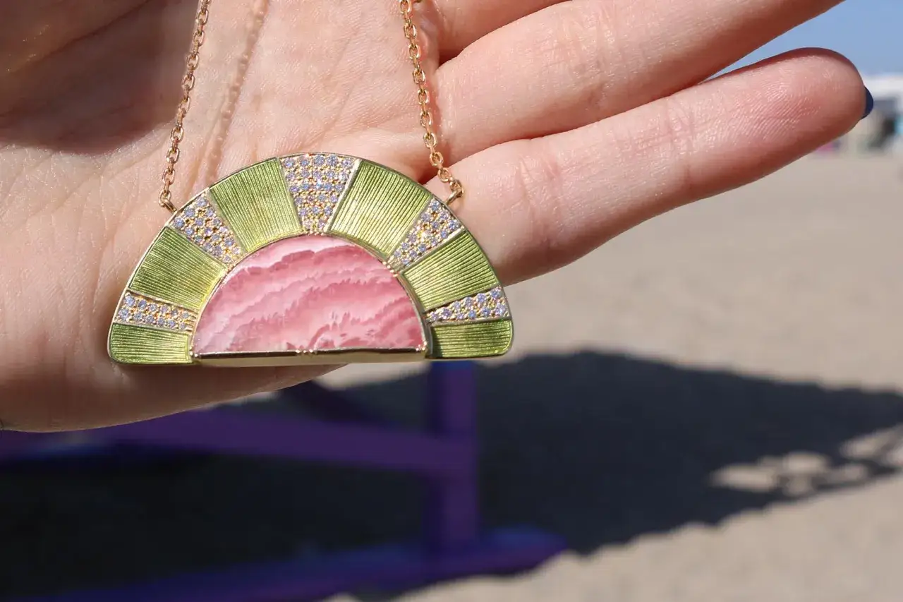 A hand holds a fan-shaped necklace with a pink rhodochrosite stone, showcasing its beauty and potential rhodochrosite benefits.